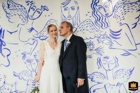 In Auch, France, a newly married bride and groom pose inside a historic chapel, standing close together against a striking and ornate wall decorated with printed angels rendered dramatically in blue ink.